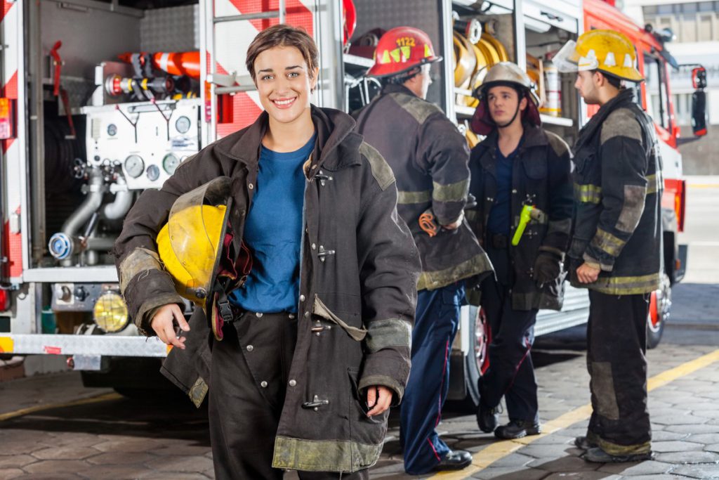 Portrait of happy firewoman with male colleagues discussing by truck in background at fire station