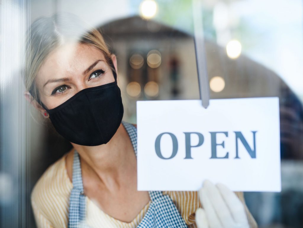 Coffee shop woman owner with face mask, open after lockdown quarantine. Shot through glass.