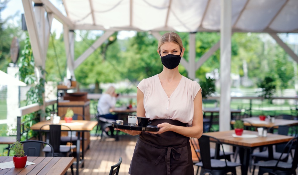 Portrait of waitress with face mask serving customers outdoors on terrace restaurant; concept: unions are standing up for workers as they go back to work