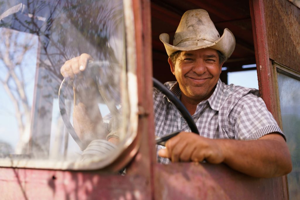Farming and cultivations in Latin America. Portrait of middle aged hispanic farmer sitting proud in his tractor at sunset, holding the steering wheel. He looks at the camera and smiles happy.