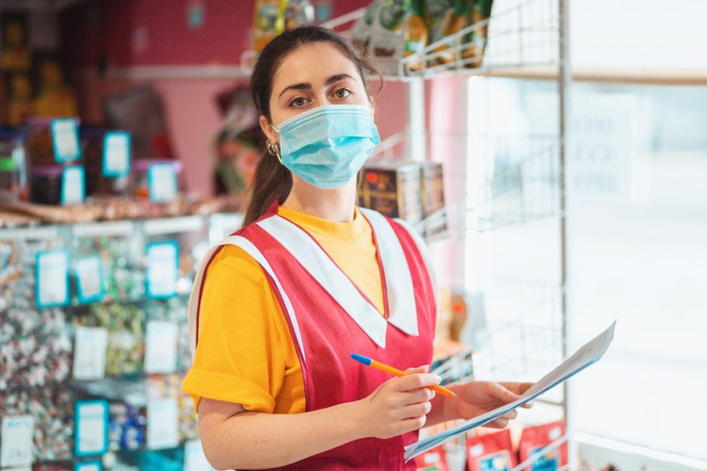 Food store. Portrait of a female employee in uniform with a medical mask on her face, holding documents. The concept of protecting against coronavirus at work.