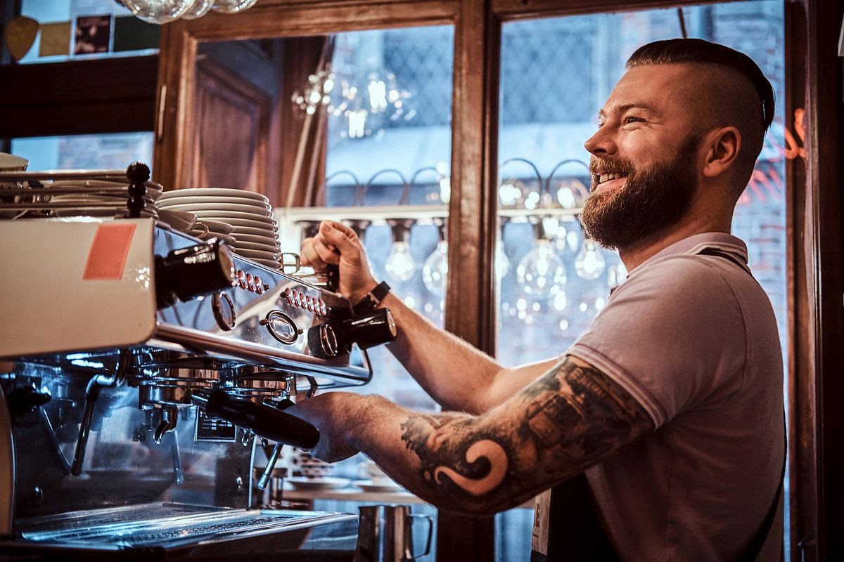 Cheerful barista with stylish beard and hairstyle making coffee for a customer in the coffee shop; union drives concept