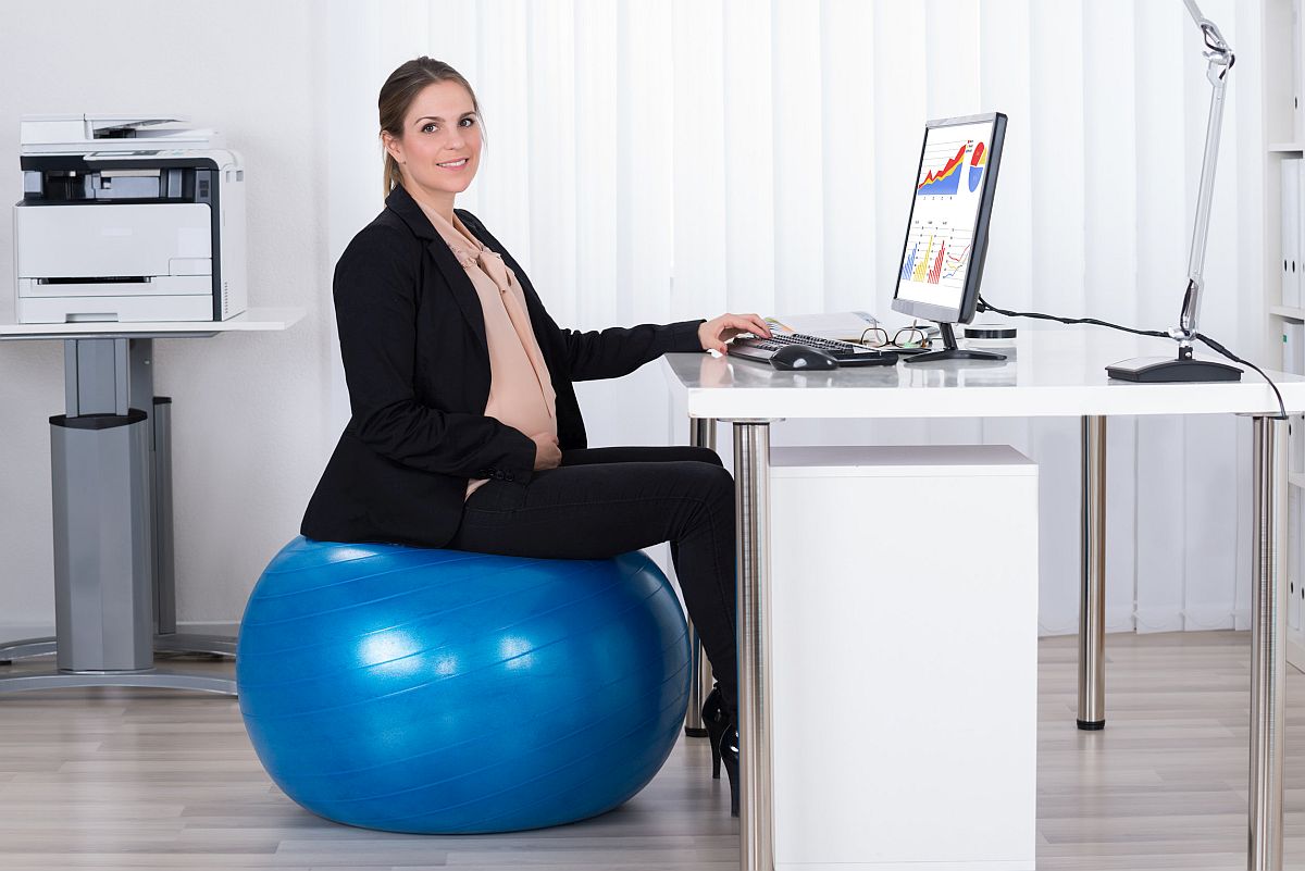 Pregnant Businesswoman Sitting On Fitness Ball While Working On Computer In Office; unions gender equity concept