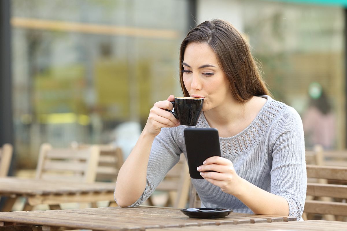 Young woman drinking a cup of coffee checking her smart phone on a coffee shop terrace; unionize using digital tools concept